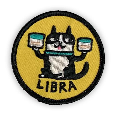 A circular embroidered patch. A black and white cat balances two fish bowls on either side. The word "Libra" sits below. The patch is yellow with a black border.
