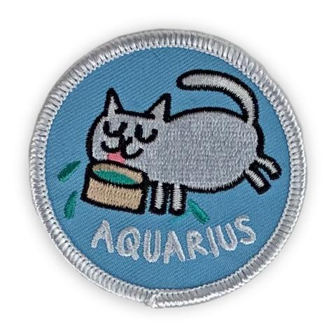 A circular embroidered patch. A white cat outlined in black laps at a bowl of water above the word "Aquarius" in white. The patch is light blue with a white border.