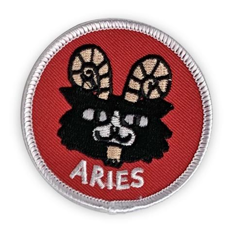 A circular embroidered patch. A ram with a cat's face is the focus. It is black with white details, and gold horns. It sit above the word "Aries" in white. The patch is red with a white border. 