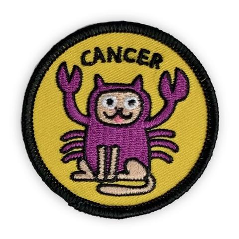 A circular embroidered patch. A tan cat is dressed as a purple crab below the word "Cancer" in a black font. The patch is yellow with a black border.