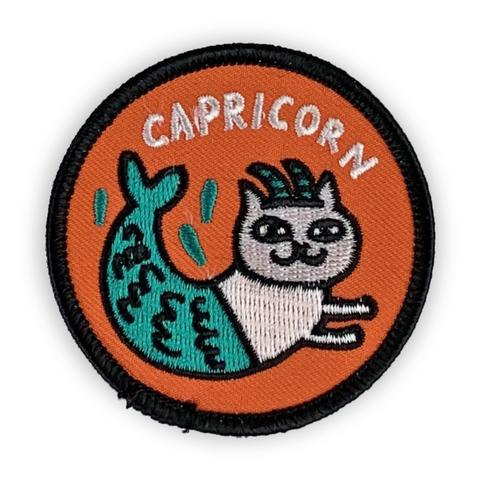 A circular embroidered patch. A white cat with a blue mermaid tail and horns is the focus. The word "Capricorn" in white font is above. The patch is orange with a black border. 