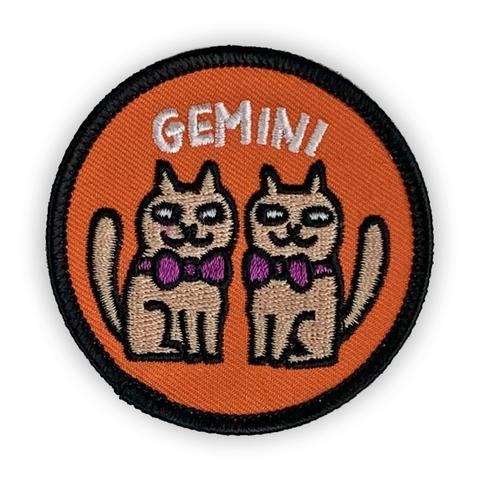 A circular embroidered patch. Two tan cats wearing purple bowties sit under the word "Gemini". The patch is orange with a black border. 