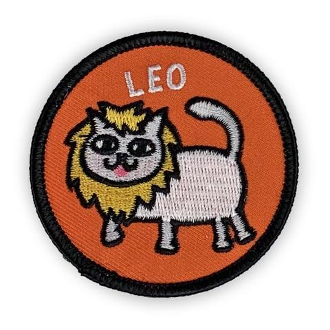A circular embroidered patch. A white cat with a yellow lions mane sticks his little tongue out. The word "Leo" sits above. The patch is orange with a black background.