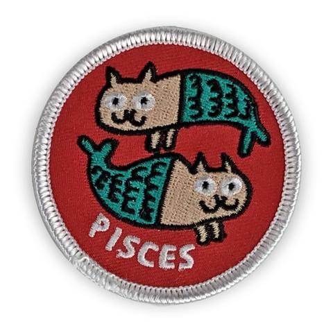 A circular embroidered patch. Two tan cats with green fish tails circle one another. The word "Pisces" sits below. The patch is red with a white border.