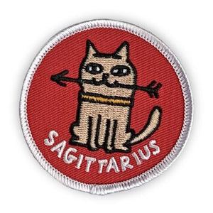 A circular embroidered patch. A tan cat wearing a yellow collar holds an arrow in its mouth. The word "Sagittarius" sits below. The patch is red with a white border.