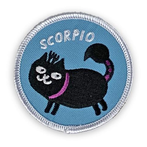 A circular embroidered patch. A black cat with a black and purple scorpion tail and forked horns is the focus. Above the cat is the word "Scorpio" in white. The patch is light blue with a white border.