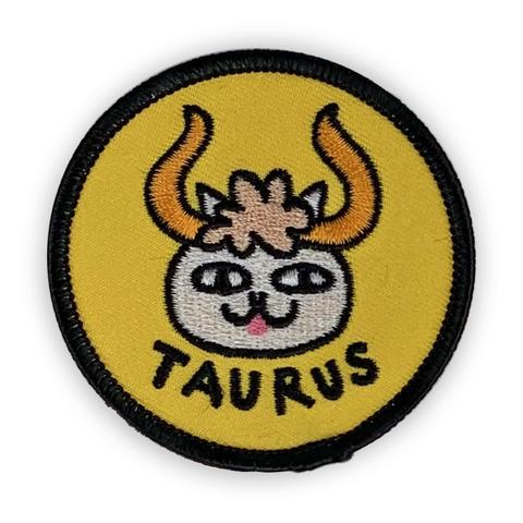 A circular embroidered patch. A white cat face with large gold horns is the focus. The word "Taurus" in black sits below. The patch is yellow with a black border.