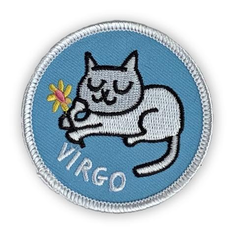 A circular embroidered patch. A white cat sits demurely and holds a pink and yellow flower. The word "Virgo" sits below. The patch is light blue with a white border.