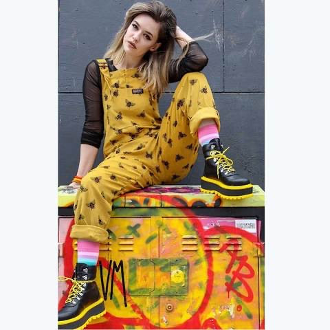 A model sits outside on a graffitied fixture wearing a pair of golden yellow corduroy dungaree overalls with an all-over honey bee pattern.