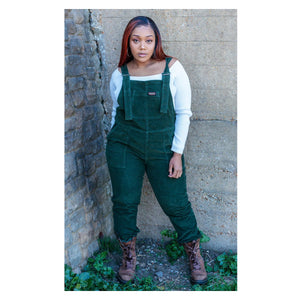 A model stands against a concrete backdrop wearing a comfortable pair of forest green stretch corduroy dungaree overalls.