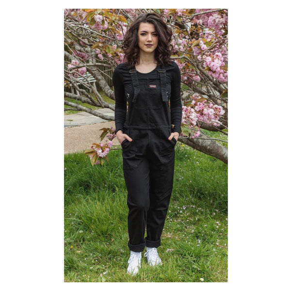 A model stands against an outdoorsy backdrop wearing a pair of comfortable plain black stretch denim dungarees.