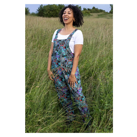 A model standing against a natural outdoorsy backdrop, wearing a pair of grey twill dungarees with a vibrant teal, pink, and orange floral pattern and light blue dinosaur silhouettes.